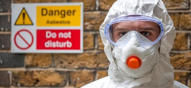 Worker in a white protective suit and respirator next to an asbestos danger sign