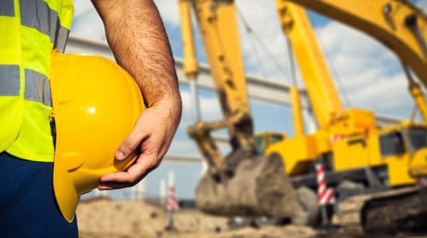 Construction worker holding a yellow hard hat beside an excavator on a Texas job site